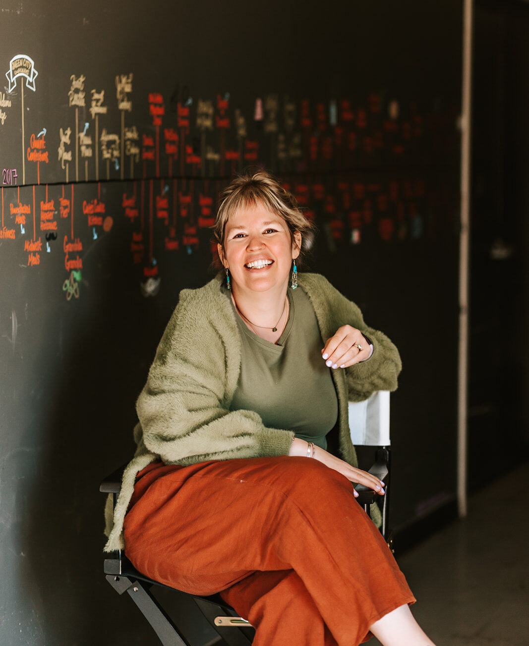 Profile shot of Rachel DesRochers, sitting in a director's folding chair in front of a chalk mural.